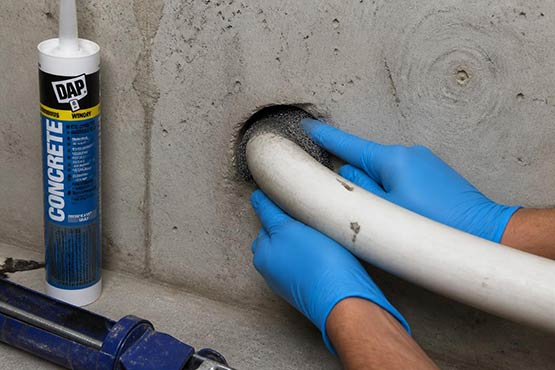Gloved hands packing stainless steel wool into a gap around a basement pipe penetration